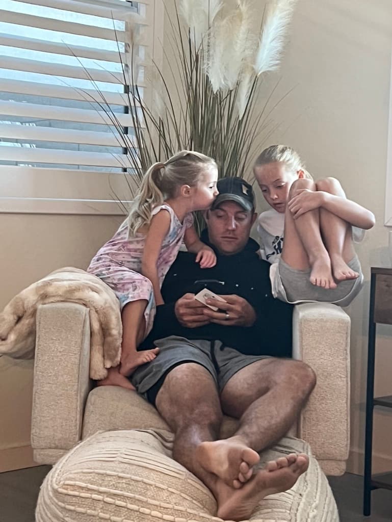Brian on a chair looking at his phone while his two daughters climb on him — one kissing his cheek, the other watching the screen